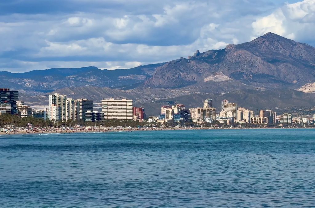 A stunning coastline features modern architecture alongside majestic mountains under a vibrant blue sky